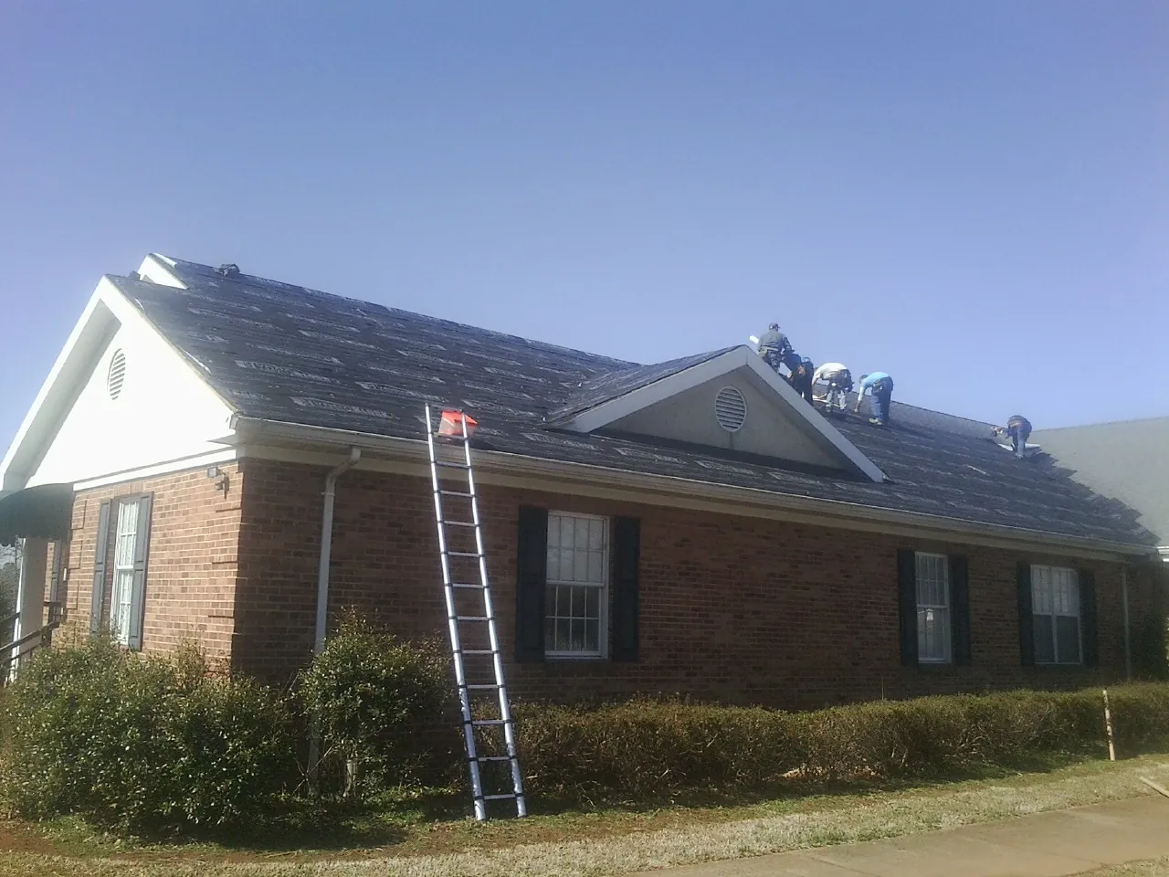Crew tearing shingles off a brick ranch roof with a ladder set — Emergency Roof Repair work by our crew in Nakina