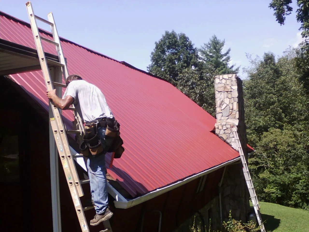 Roofer on a ladder beside a red metal roof and stone chimney — Soffit & Fascia Repair by Roofing Brothers in Nakina